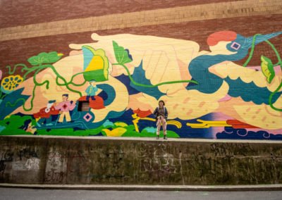 Photo of artist Wenting Li standing in front of her mural in Chinatown. It has three giant cranes intertwined with lotus flowers and roots, and carrying multiple human figures. It has colours of yellow, green, teal, red and pink.