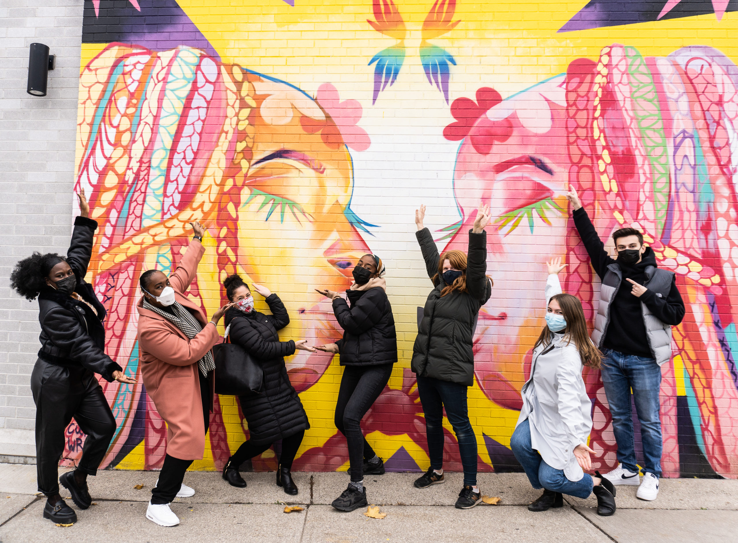 Photograph of Curtia Wright’s ‘Celebrating Queer Black Lives’ mural painted on the side of a building. The mural has two side-facing portraits, with floral patterns and is brightly coloured. There are seven people striking playful poses in front of it.