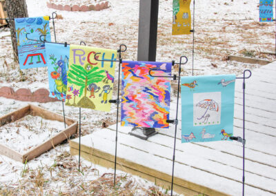 Weaving Our Roots flag garden on a snowy day. There are colourful designs created by the community with green, yellow and teal colours used throughout.