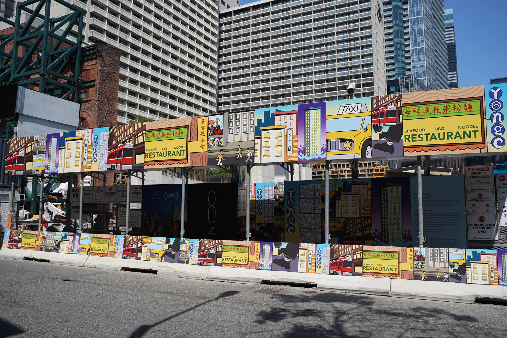 Colorful construction hoarding on an urban street features illustrated storefronts and signs with a taxi graphic.