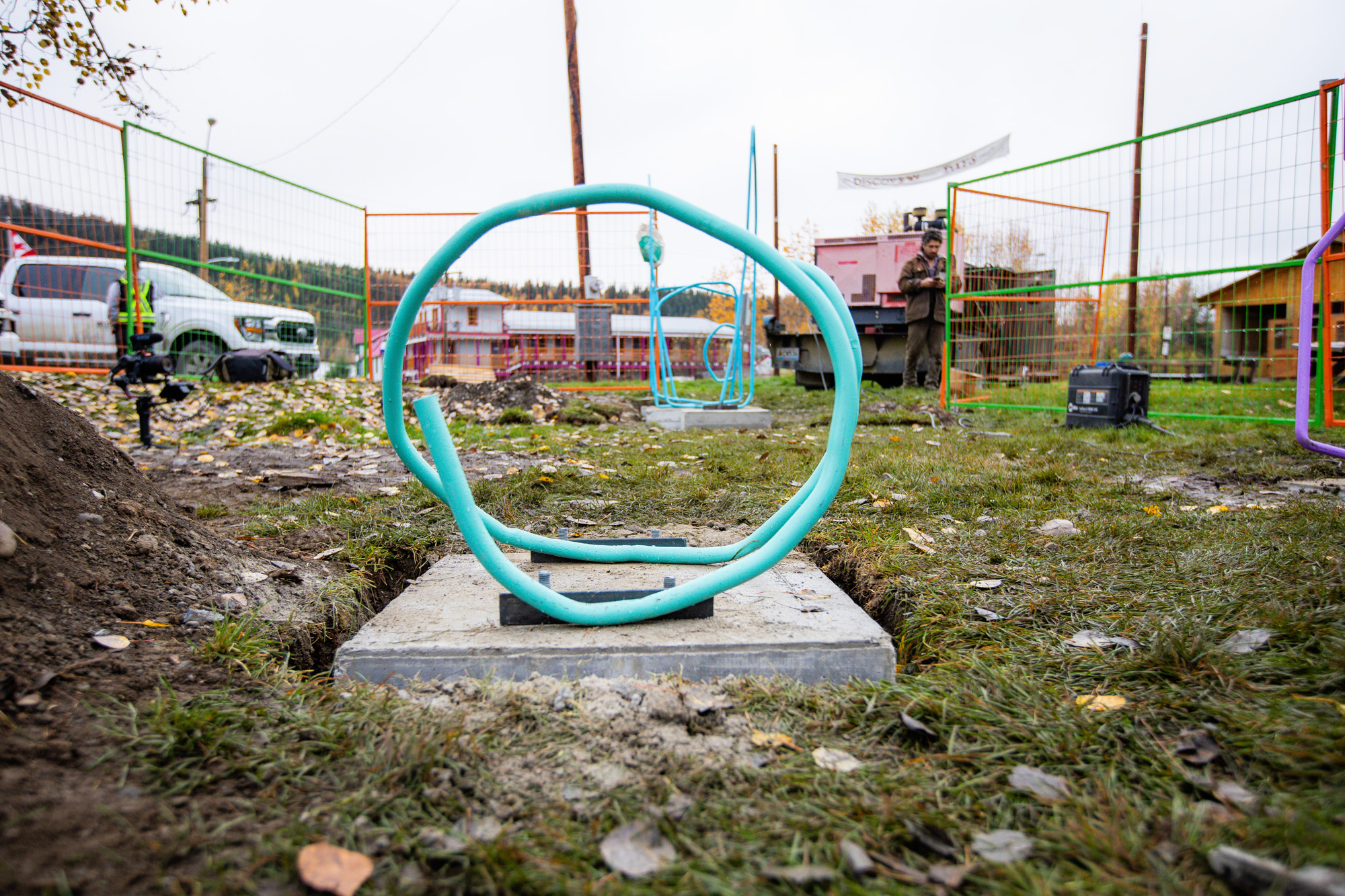 A park with a colourful, looping sculpture installed on a cement base. The ground is grassy with scattered leaves, and there are various sculptures and fenced areas in the background.