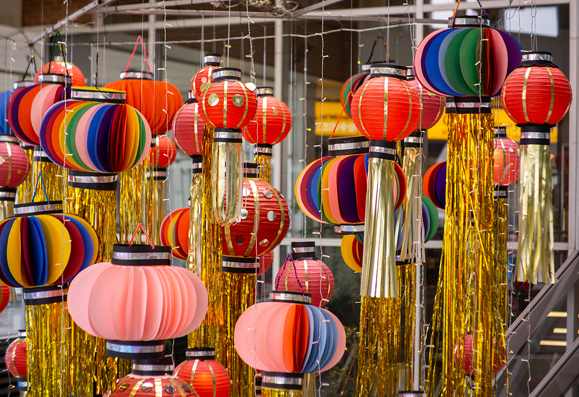 Photo of brightly coloured paper lanterns with hanging golden tassels. The lanterns are in different sizes, interspersed with fairy lights and are hanging from metal pipes.