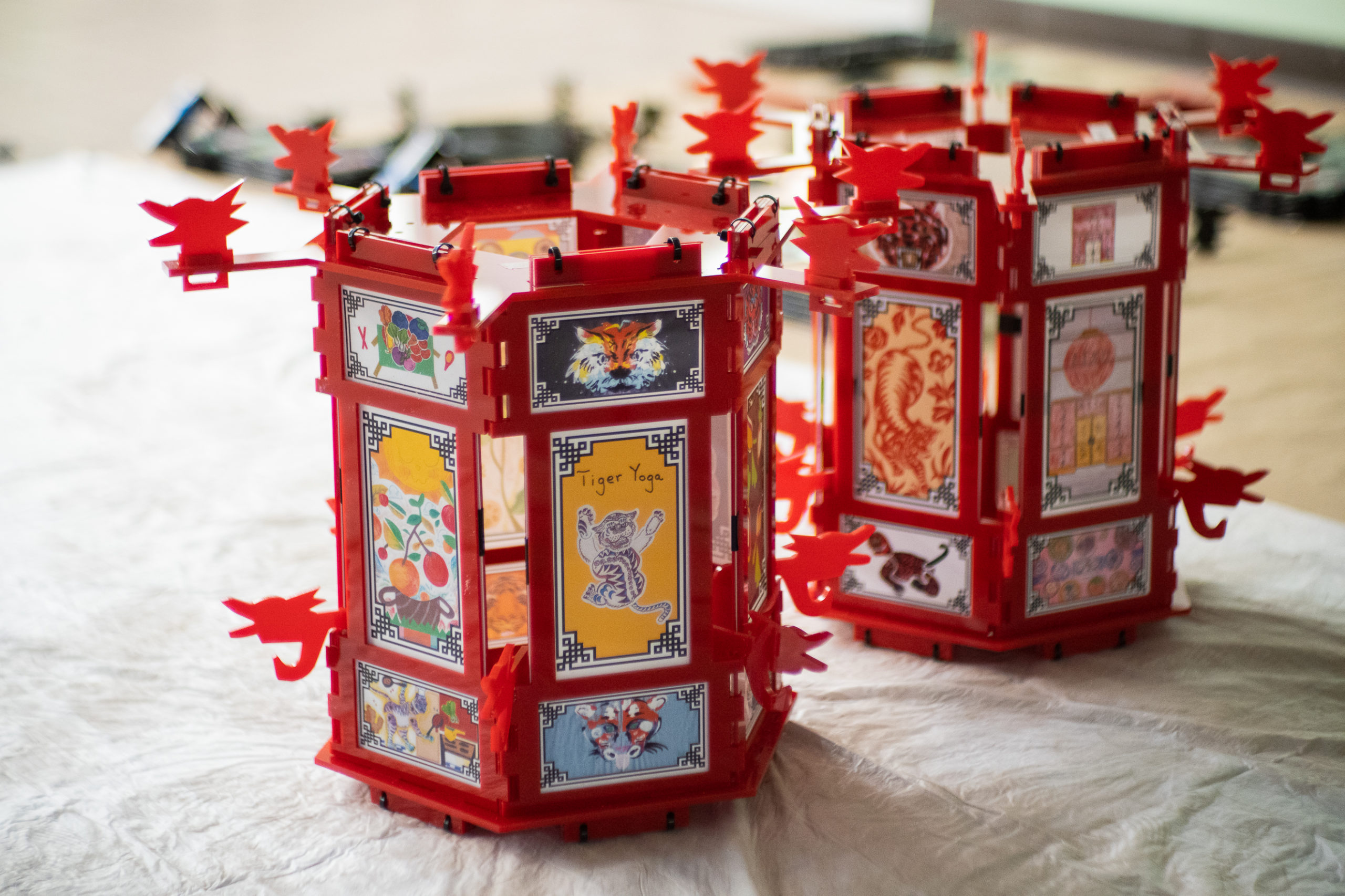 Photo of two red palace lanterns from the Yue Moon exhibit, sitting on the floor. The lantern holds different artwork featuring Lunar New Year imagery of foods and tigers.