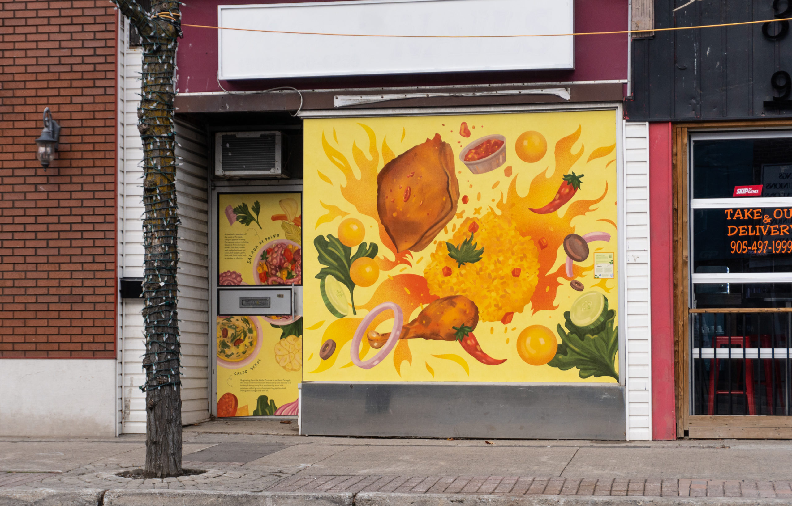 Photo of a Brampton storefront featuring the artwork of Meegan Lim, the artwork shows a Portuguese chicken feast with rice, potatoes, kale and more. Parts of the sidewalk, building and road are visible.