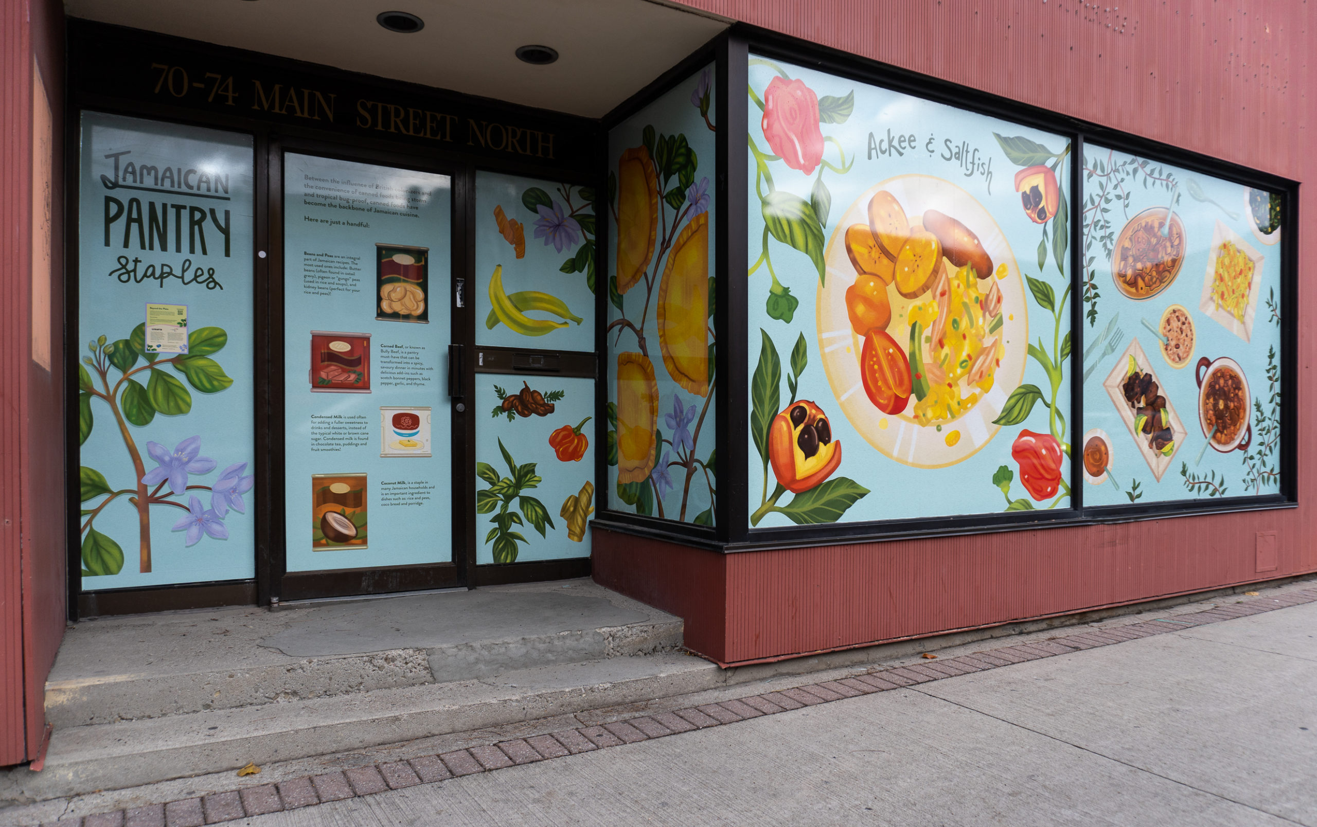 Photo of a Brampton Storefront with red wood panelling, featuring artwork by Meegan Lim. It showcases Jamaican foods on a light blue background. Part of the side walk is visible.