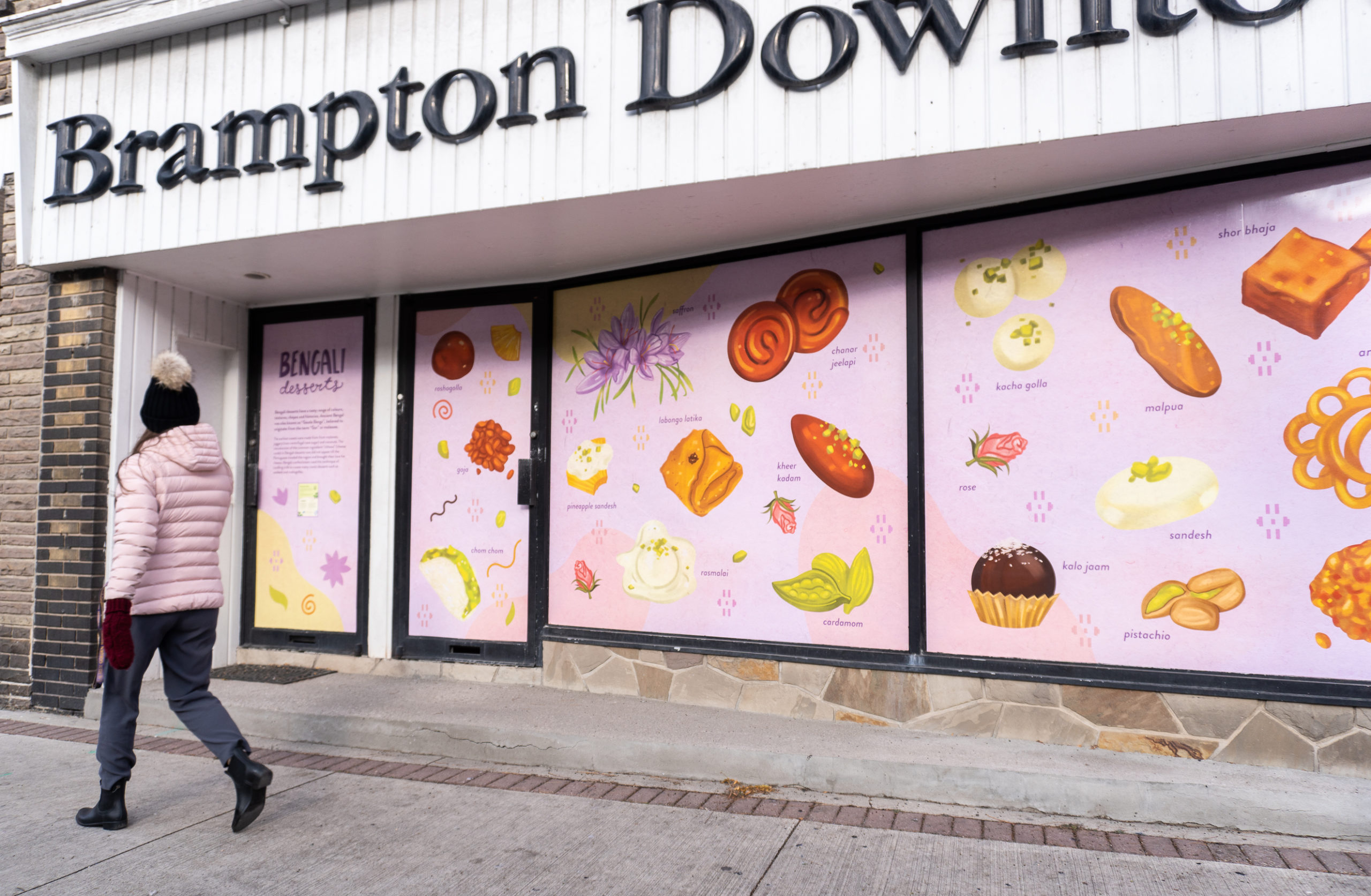 Photo of a person walking past a Brampton storefront featuring the artwork of Meegan Lim that shows different Bengali sweets on a lilac purple background.