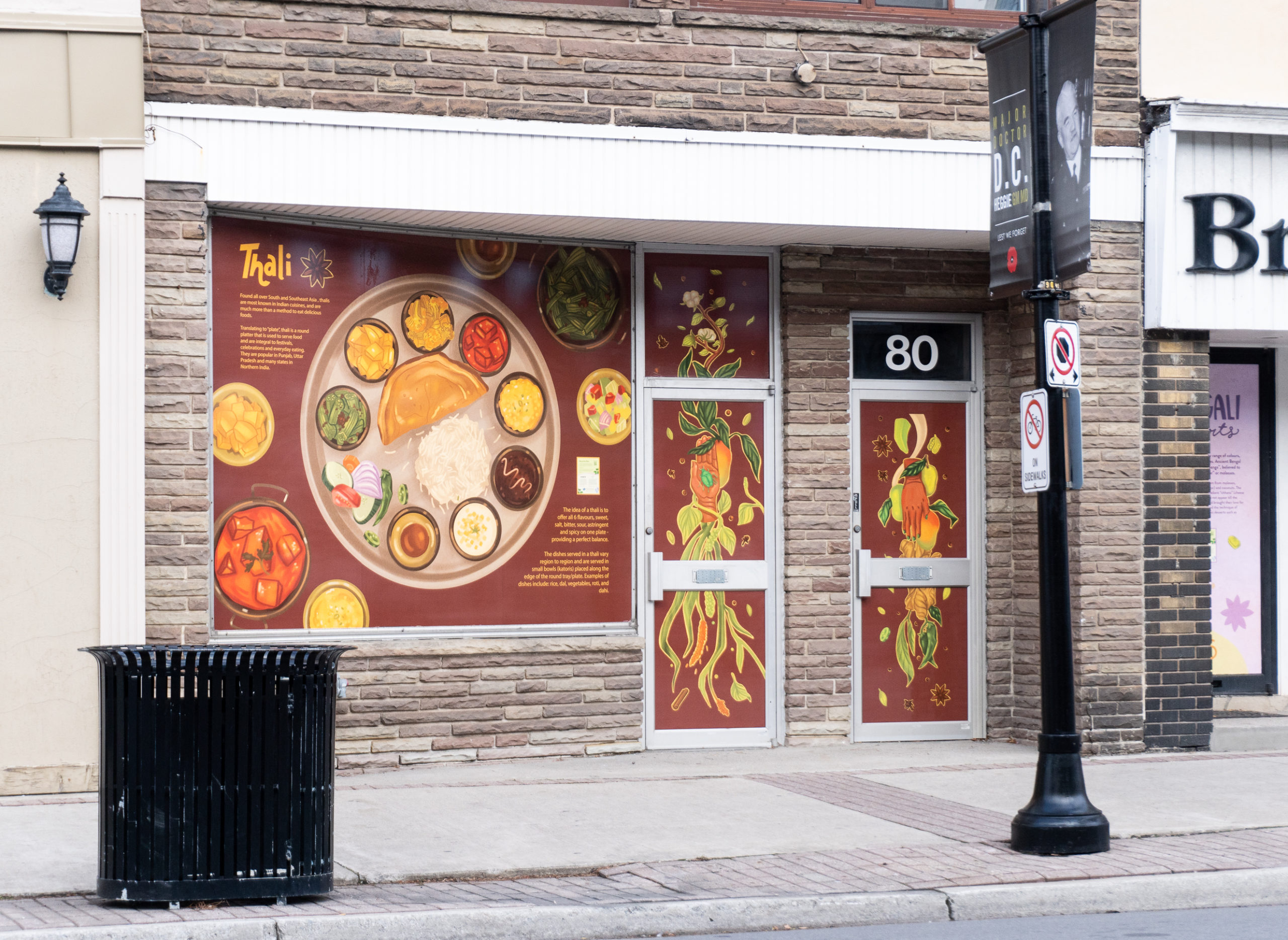 Photo of Brampton storefront featuring artwork by Meegan LIm, showcasing a Punjabi Thali meal. Parts of the sidewalk, buildings and road are visible.