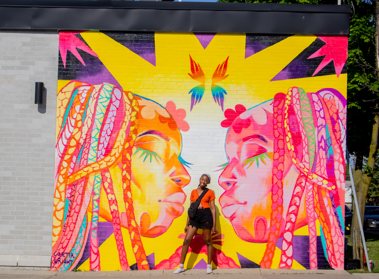 Artist Curtia Wright standing in the very centre of their Celebrating Queer Black Lives mural, painted on the wall of a grey and black coloured Beer Store. The mural has two nearly identical feminized faces in pastel colours yellow, pink, blue, and purple, and these faces are facing each other, with a rainbow coloured butterfly featured above the faces at the top centre. Curtia is wearing white running shoes, black shorts, and an orange T-shirt.
