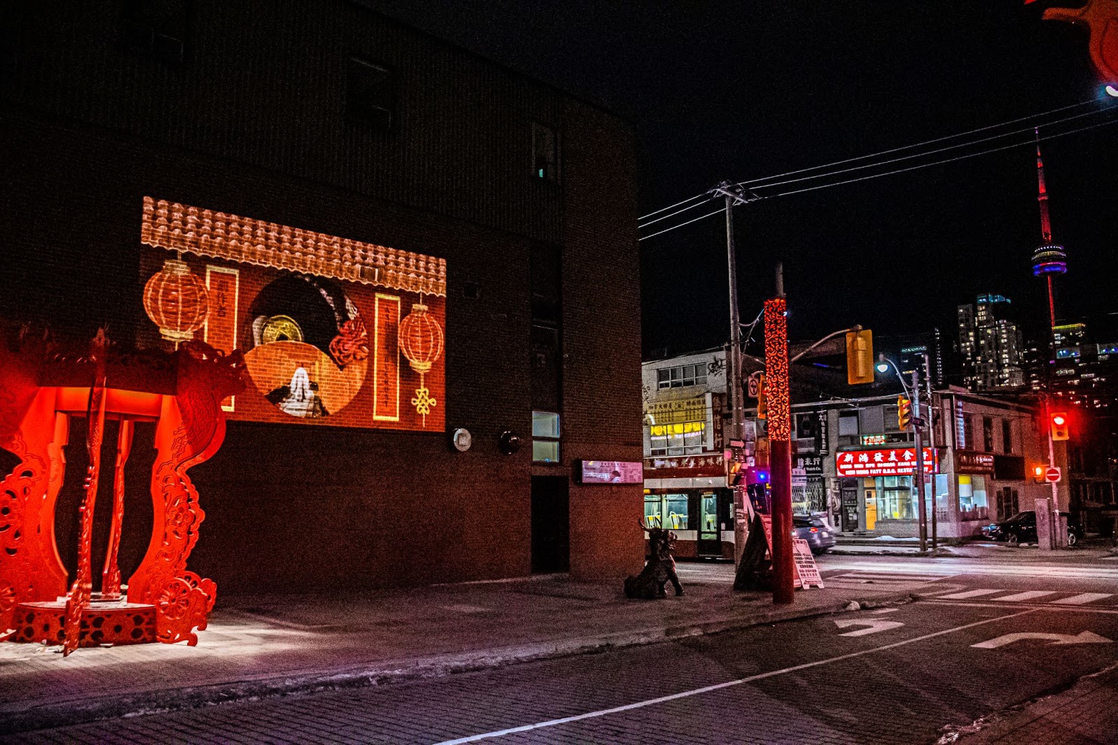 Snapshot of Toronto's Chinatown on a cold winter night. The CN Tower is lit up with rainbow colours faintly visible in the background. The Animated Light stop-motion feature, seen on the left on the side of Chinese Gospel Church, lights up the streets with bright and vivid shades of red for Lunar New Year. 