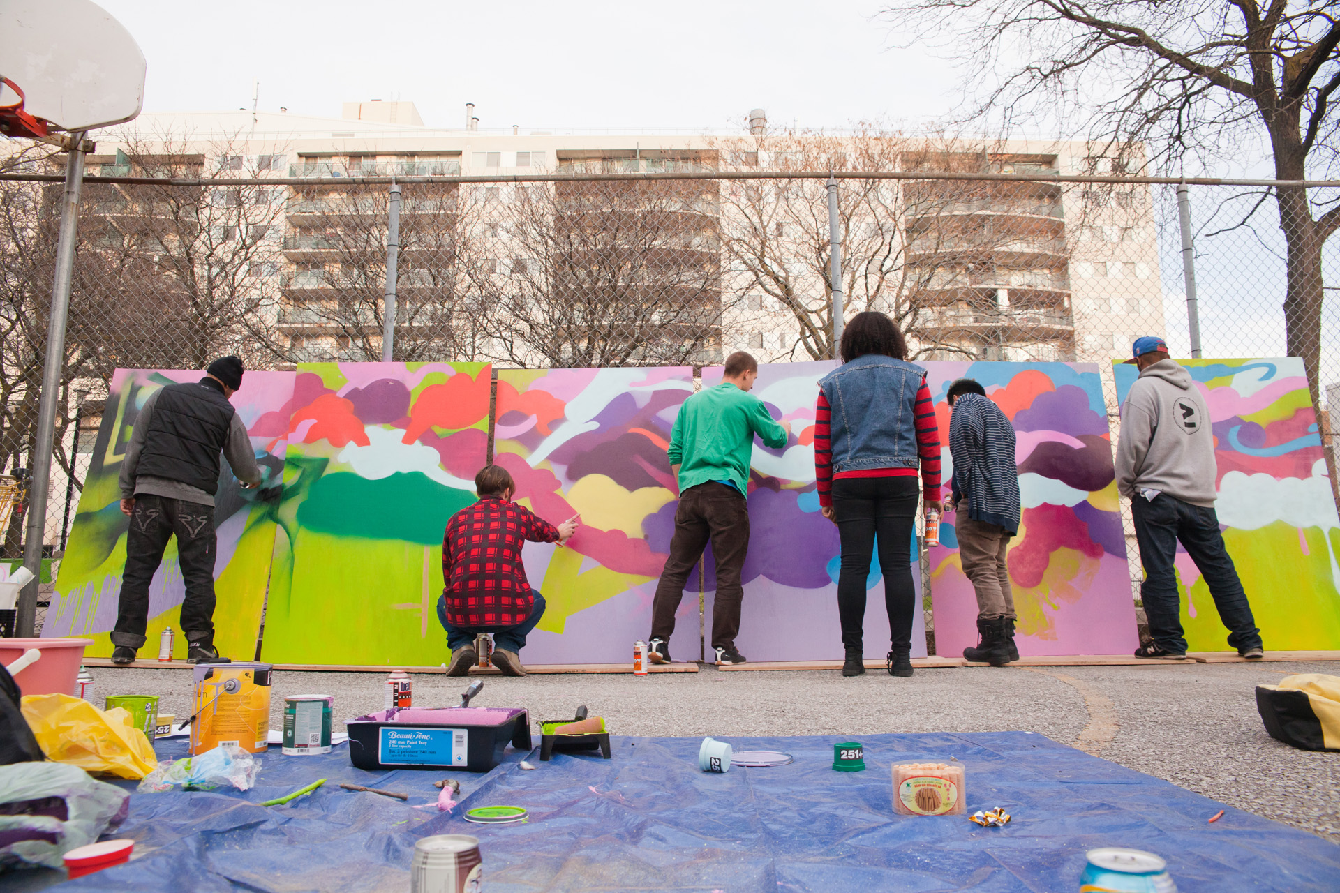 A group of people painting outdoors on a long standing panel. An apartment building is in the distance.