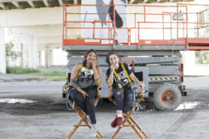 Two mural artists sitting outside under the Toronto Gardiner highway and smiling at the camera