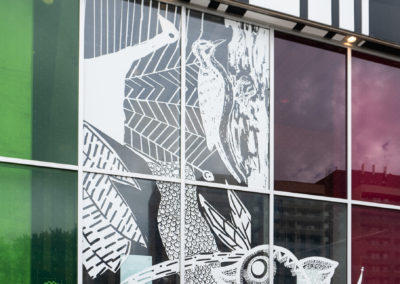 Photo of Safe Flight Home white vinyl window installation, featuring drawings of birds. The sit has several panes of glass some that are clear and others tinted green and pink. The top of the building that has vertical black and white stripes, and cement sidewalk is visible.