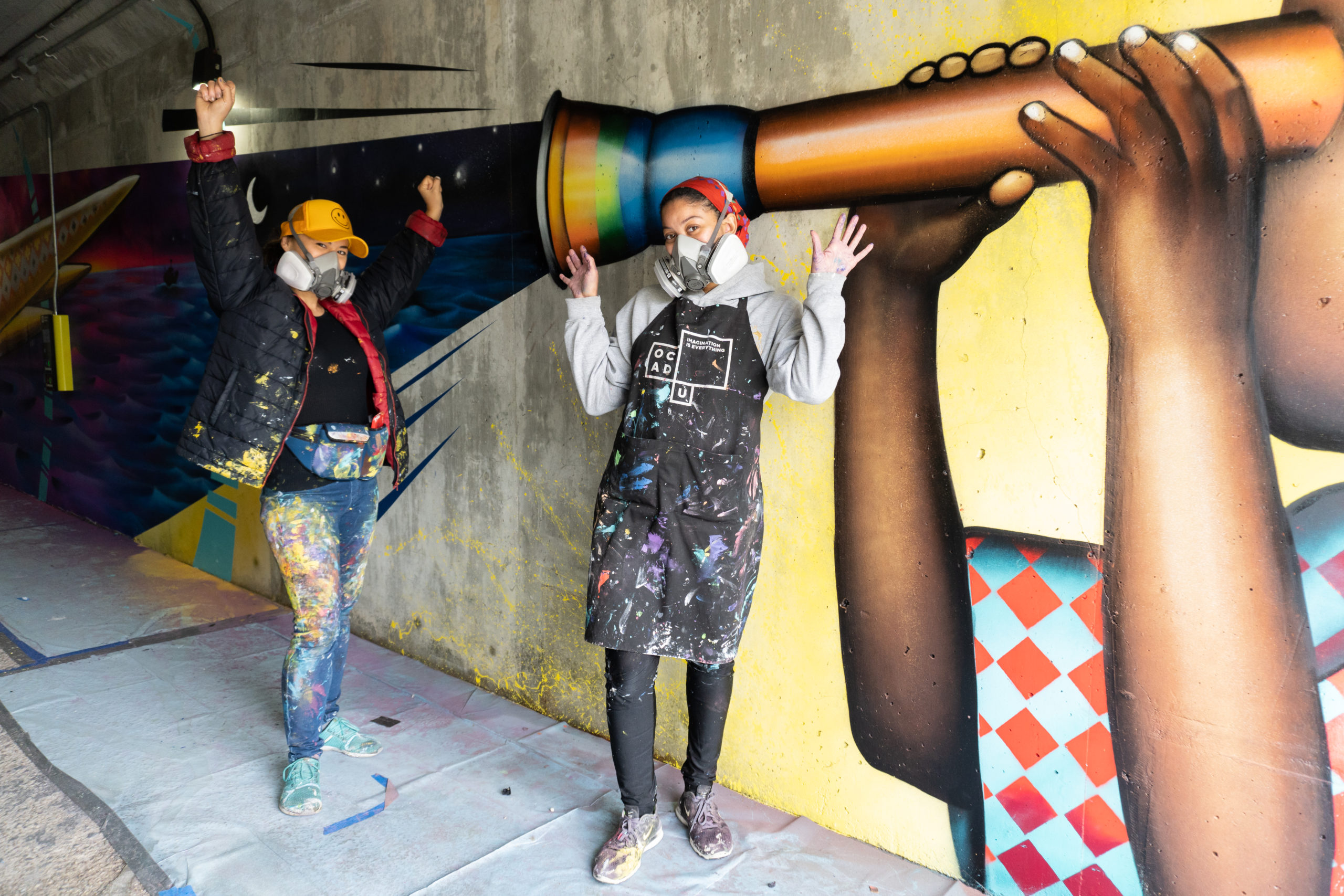 Photo of two mural Assistants (Edan Maxam and Kseniya Tsoy), standing in front of the mural inside of Wilson TTC Station of a boy holding a telescope. They are both holding their arms up in a pose as if they are holding the telescope mural.