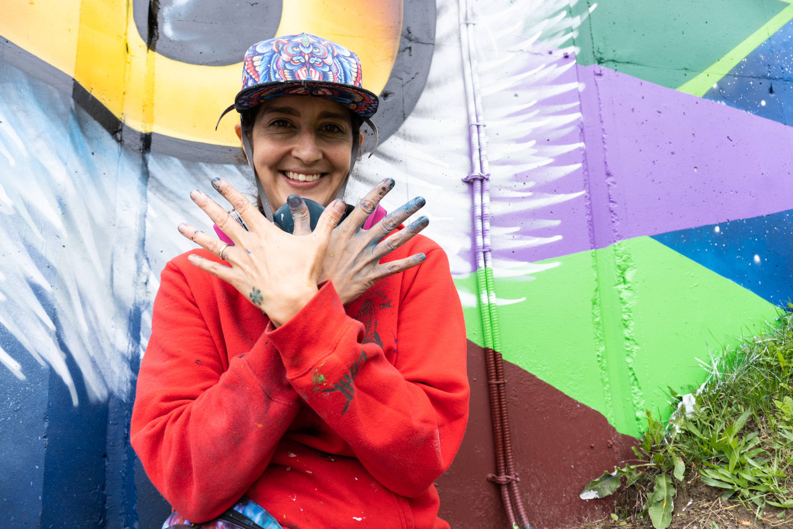 Photo of artist Shalak Attack wearing a red hoodie and hat, holding up her hands making a wing/bird shape. She is standing in front of a colour mural with colours of yellow, purple, green, blue and burgundy.