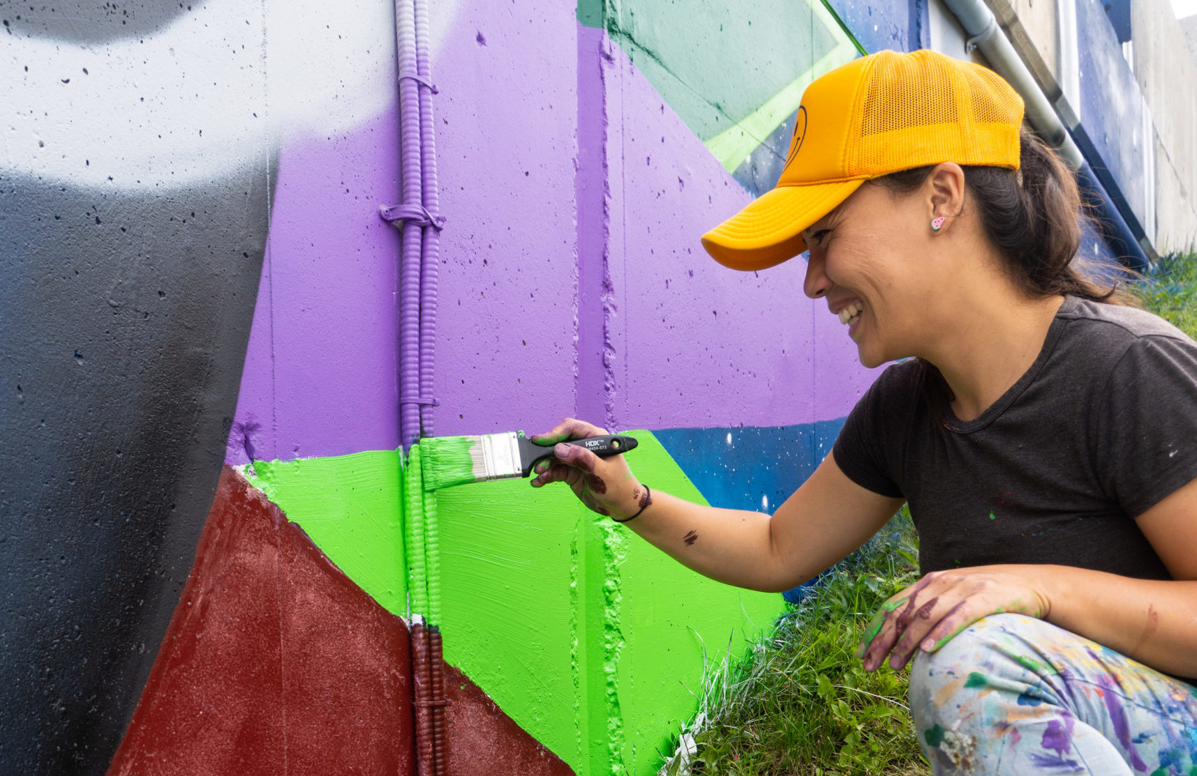 Cropped landscape photograph of art assistant Kseniya Tsoy painting the mural outside of the Wilson TTC Station entrance. She is smiling, wearing a yellow hat, grey tee and jeans. The mural has geometric shapes in colours of purple, green, blue and red.