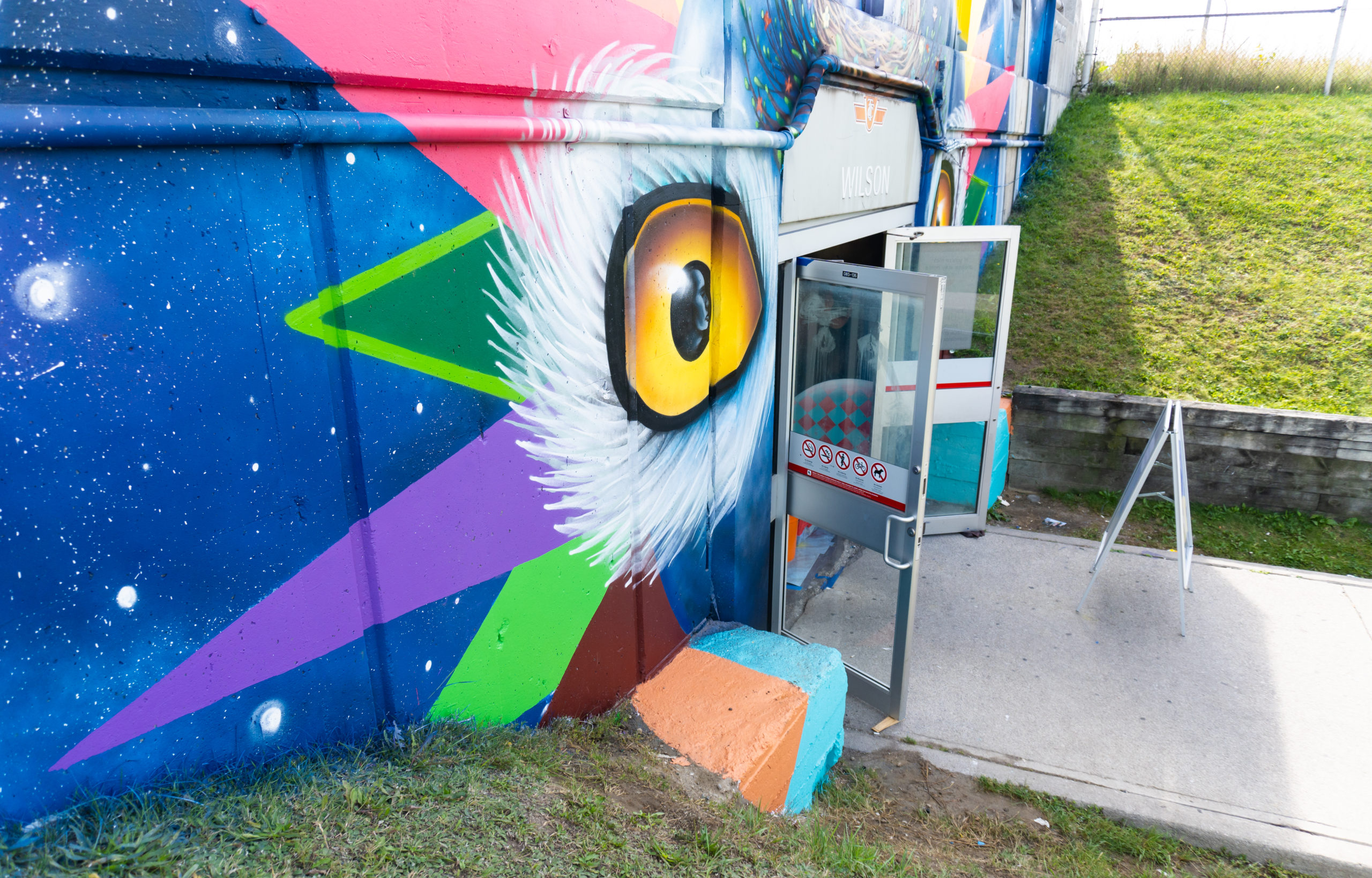Photo of mural by Shalak Attack at the Tippett Rd entrance, doors are open and the photo is taken from an incline on the left side. The mural features the eyes of a snowy owl, the earth, and geometric colourful shapes.