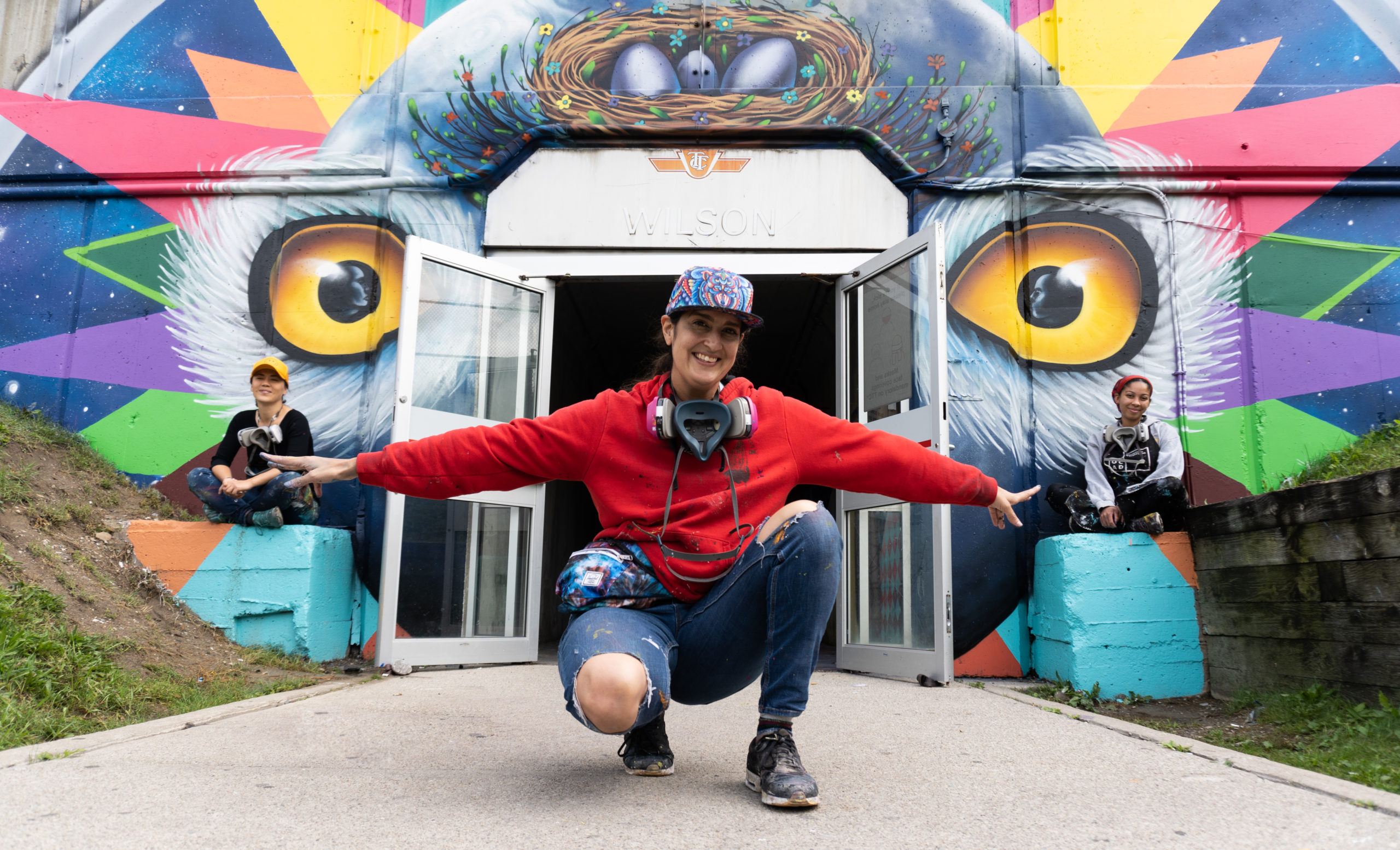 Photograph of artist Shalak Attack, mural assistants Edan Maxam and Kseniya Tsoy posing in front of their mural at Wilson TTC station entrance. The mural is brightly coloured, featuring a bird with geometric shapes and golden eyes. Shalak Attack is closest to the camera, kneeling with arms spread out, pointing to Edan and Kseniya on either side of the station doors sitting on 2 concrete pedestals.