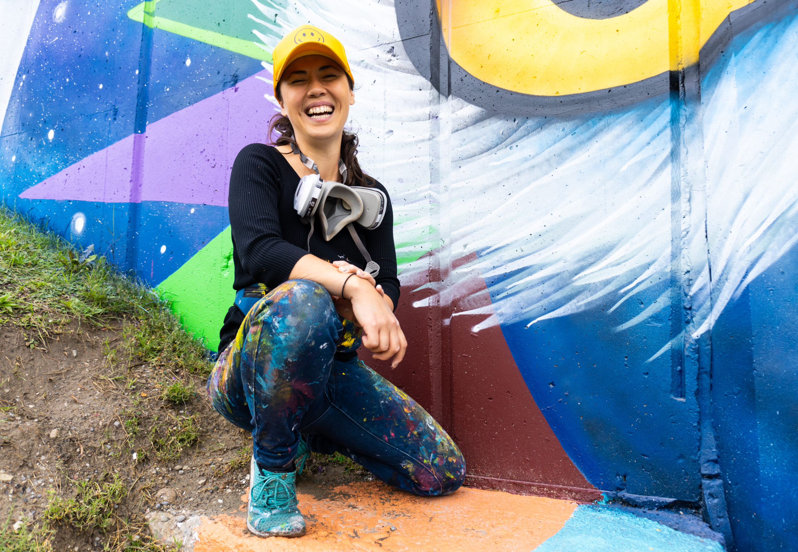 Photo of mural assistant Kseniya Tsoy, she is smiling at the camera, wearing a yellow hat, black shirt, jeans with paint on them and teal shoes. She is kneeling in front of a mural that has an owl eye with colours of blue, yellow, purple, green and burgundy.