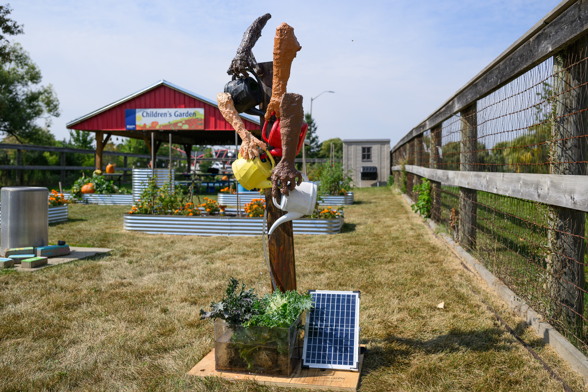 A whimsical garden sculpture of a person made from tree branches holding a watering can stands on grass beside solar panels. In the background, a red-roofed structure labeled Childrens Garden is visible.