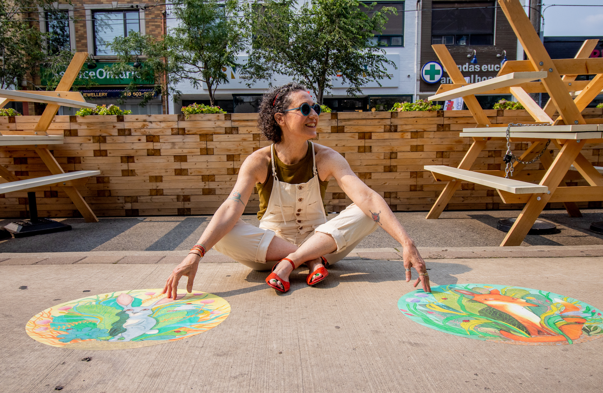 Yaara Eshet sitting on the ground beside large, round sidewalk decals that they designed. The designs have illustrated foxes and rabbits.