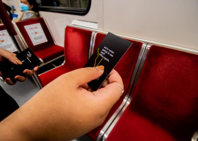 Photo of two hands, one holding a single black card with white text "we're never going to see each other again" and the other holding another stack. The background has 5 red TTC subway seats