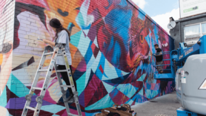A large colourful mural being painted by a female artist standing on a ladder