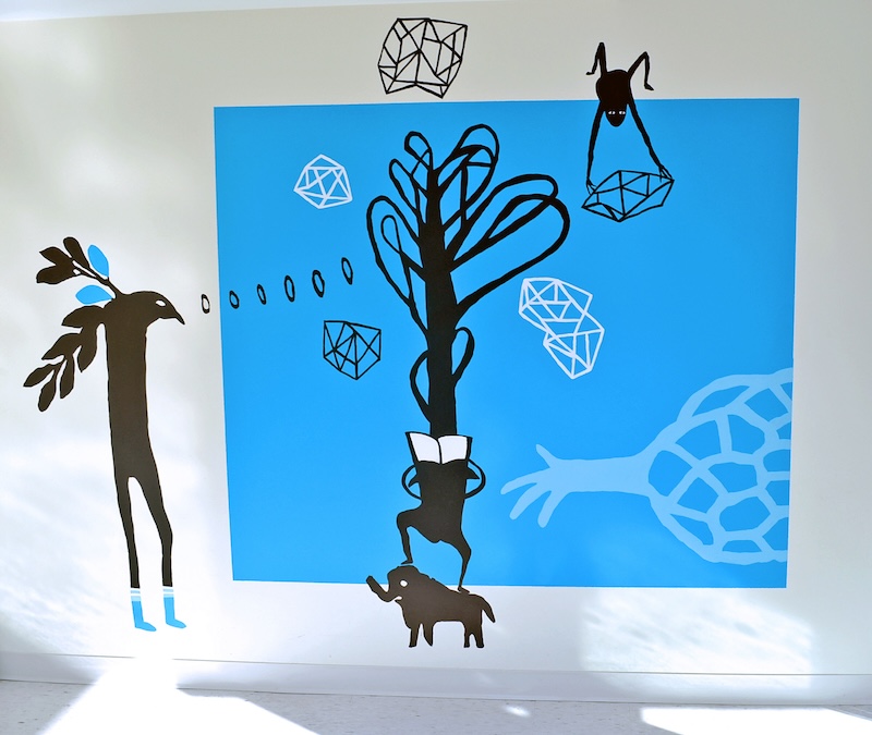 An indoor mural on a white wall in Art Hub 27 by Z'otz* Collective, There is a blue square with black and white illustrations