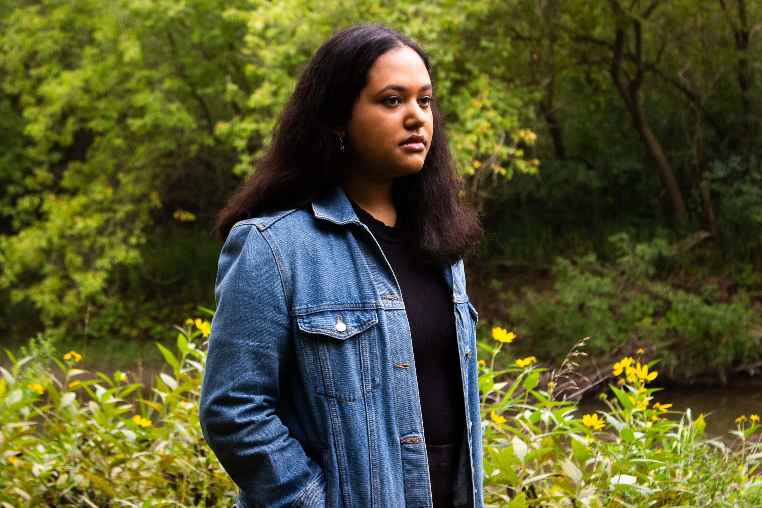 Zara Rahman in Rowntree Mills Park. Zara has medium length, dark hair and is wearing a blue button up shirt and a black t-shirt.