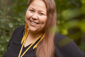 Lindsey Lickers with long brown hair and wearing a black shirt and yellow accessories. Lindsey is smiling at the camera.