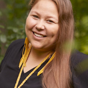 Lindsey Lickers with long brown hair and wearing a black shirt and yellow accessories. Lindsey is smiling at the camera.