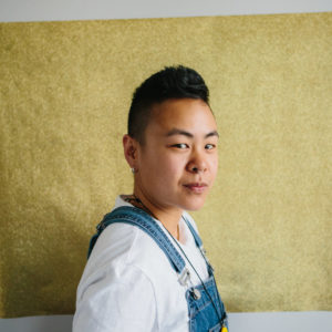 Headshot of Paddy Leung (PUFF Paddy) who is Asian Canadian with short hair. They are wearing a white t-shirt and blue overalls and standing against a gold, textured backdrop
