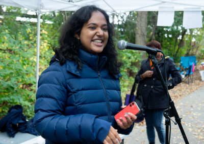 Photo of spoken word artist Zara Rahman wearing a blue puffer jacket and holding a phone, at a microphone in a park.