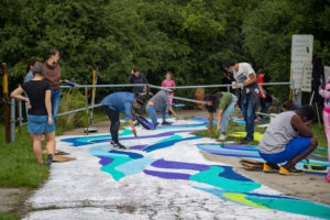 Photo of people outdoors in the Rowntree Mills Park, painting a colourful ground mural with colours of teal, blue, purple and green. There are trees and lush foliage in the background.