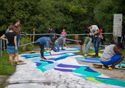 Photo of people outdoors in the Rowntree Mills Park, painting a colourful ground mural with colours of teal, blue, purple and green. There are trees and lush foliage in the background.