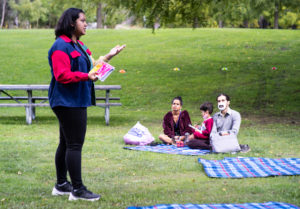 Photo of Zara Rahman performing spoken word poetry at Rowntree Mills Park, there is a family of three seated on a picnic blanket watching, hills of grass and trees in the background.