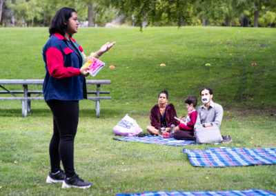 Photo of Zara Rahman performing spoken word poetry at Rowntree Mills Park, there is a family of three seated on a picnic blanket watching, hills of grass and trees in the background.