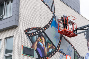 An artist on a crane high up against a building to paint a mural. The mural is in progress with rough sketches of a film strip with imagery of children taking photos with disposable cameras. The artist is working on a blue jay bird.