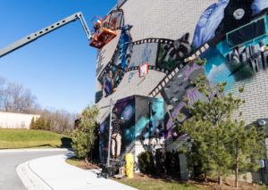 A sunny day with artists working on a mural production. One artist is spray painting a galactic scene at the bottom while the second artist is on top of a crane and working on a larger piece at the top of the building.
