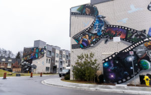 A series of large murals on the side of community homes on an overcast day. The artwork reflects movie film strips with images of animals, Black community members, and children that represent the residents of the community