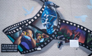 A close-up of a large public art mural on the side of a residential complex. It is a design of a film strip with each strip showcasing realistic images of nature, animals, and two Black children taking photos with disposable cameras