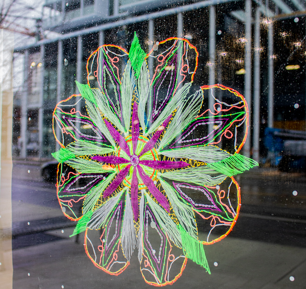 An 8-pronged mandala placed on a window; rainy background with glass building seen on the other side of window. The mandala has purple prongs on the inside and lime green prongs surrounding them, with an intricate orange pattern surrounding the design. 