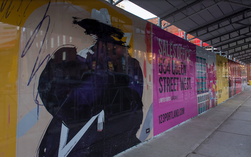 Hoarding exhibit with illustration of woman enveloped in a black blob, with yellow background; neon pink advertisement of 123portland.com next to illustration. An empty sidewalk is immediately to the right of the exhibit, with a brown building slightly visible in the far background.