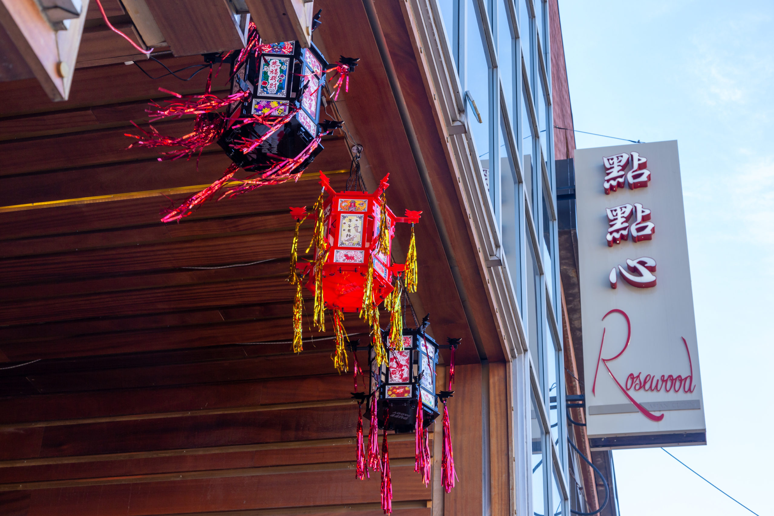 Vertical sign on the right that reads "點點心 Rosewood" in red text on a white background. Three hand-crafted lanterns, two black and one red, are seen on the left with their red and gold tassels blowing in the wind.