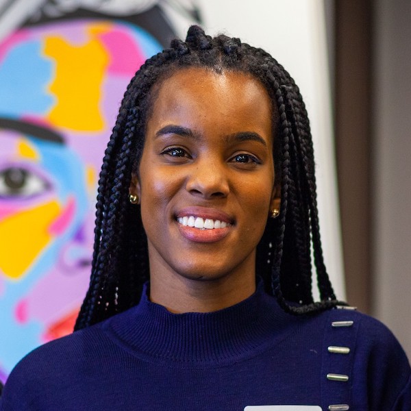 Headshot of Alica Hall in a dark sweater in front of a wall with a colourful mural of a person