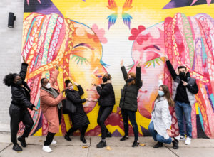 A group of people dressed warmly and in face masks celebrating and pointing at a large mural of queer Black femmes