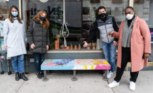 Four people dressed in warm winter clothing and face masks posing beside a colourfully painted bench in front of a storefront