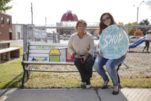 Artists Aggie Armstrong and Rhonda Franks sitting on a painted outdoor bench and holding a sidewalk decal created by the artist for Downtown Tillsonburg
