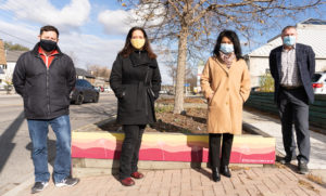 Four people wearing face masks and posing in front of a pink and yellow painted pollinator box on a sunny day