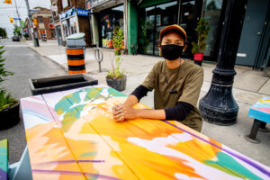 Leone McComas wearing a black face mask and sitting at a picnic table that she painted in pastel orange, green and yellow
