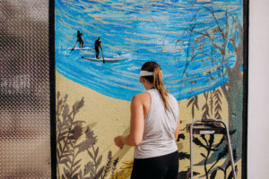 Jocelyn Kearns painting a large outdoor mural. The mural features figures paddle boating and a beach
